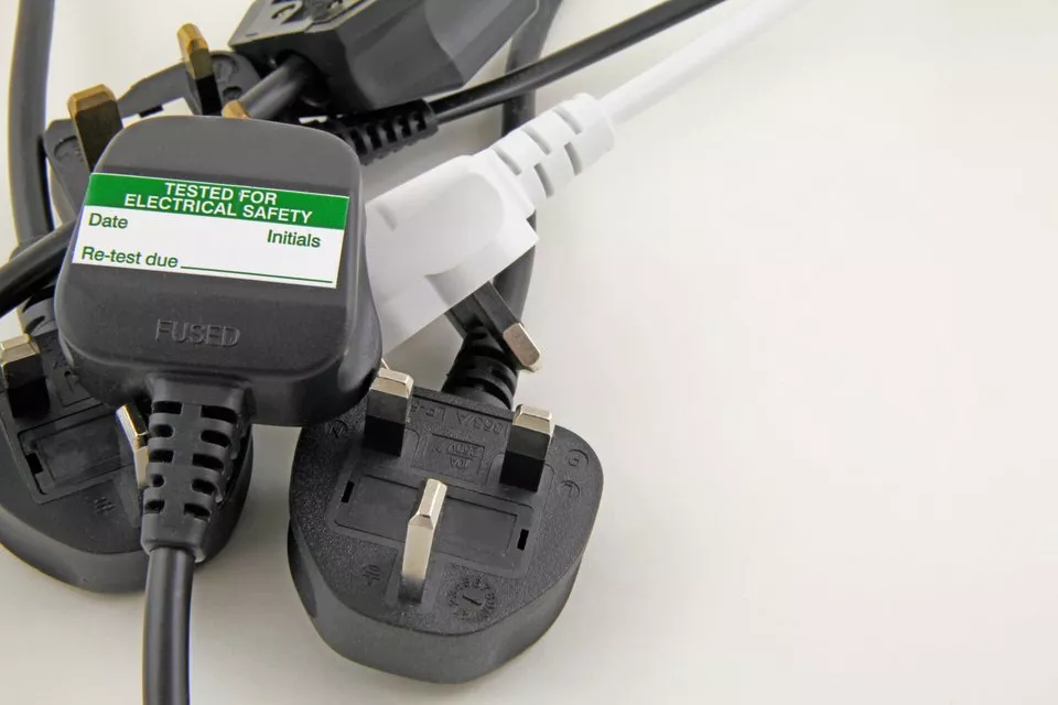 A black plug in a white wall socket on a grey wall, illustrating electrical safety in PAT testing.